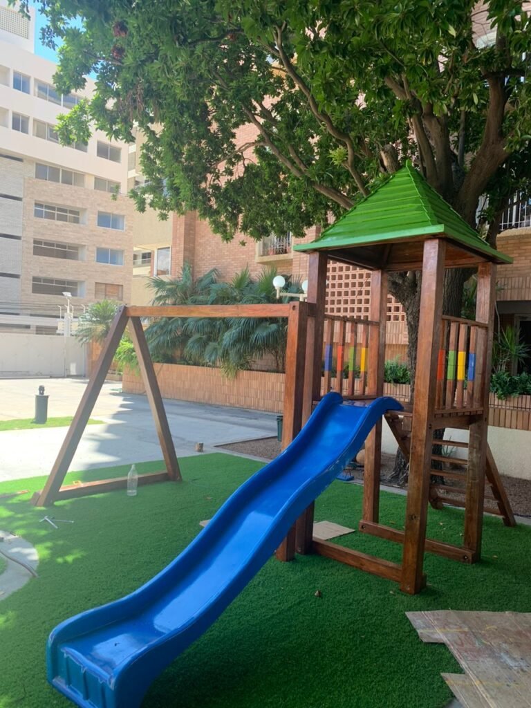 Torre de juegos en madera con tobogán azul instalada en el parque infantil del Edificio Luna